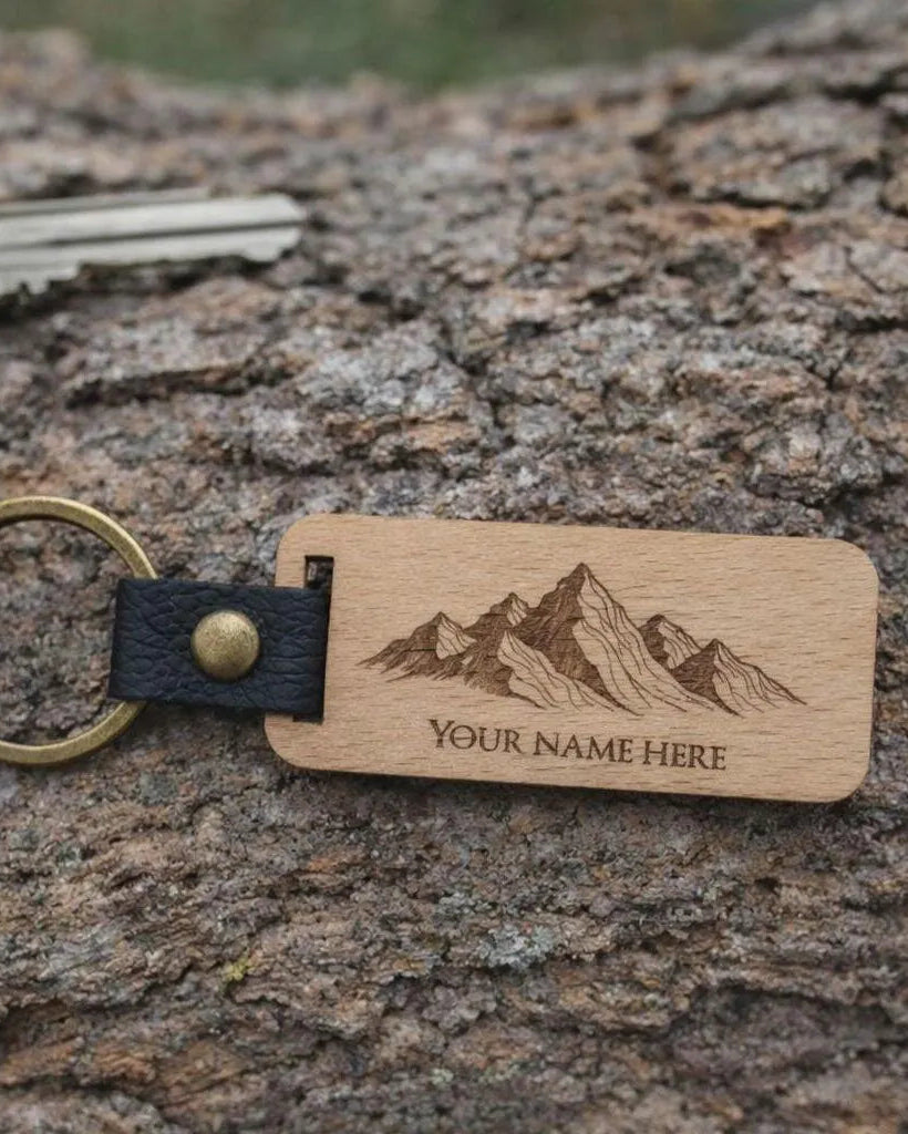 A personalized wooden key tag with a mountain engraving and the text 'YOUR NAME HERE', attached to a brass key ring and black leather loop, resting on a textured tree bark surface.