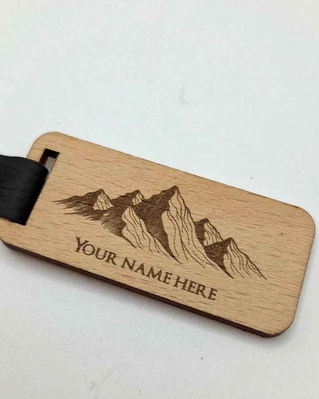 A personalized wooden luggage tag with a mountain engraving and the text 'YOUR NAME HERE', attached to a black leather strap with a brass snap, shown on a plain light background.
