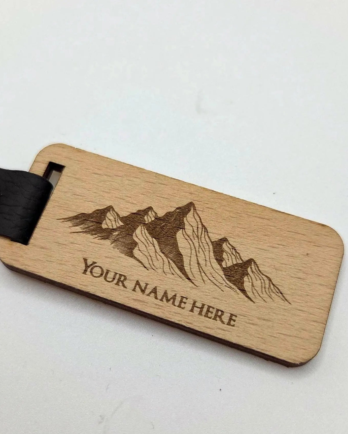 A personalized wooden luggage tag with a mountain engraving and the text 'YOUR NAME HERE', attached to a black leather strap with a brass snap, shown on a plain light background.