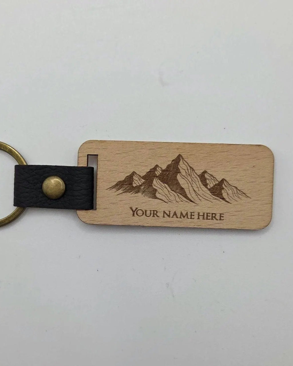A personalized wooden keychain with a rectangular natural wood tag engraved with a mountain graphic and the text 'YOUR NAME HERE'. It features a black faux-leather strap with a brass snap button and a brass key ring, shown on a plain light background.