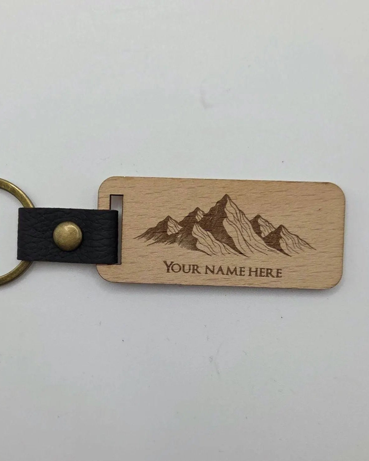 A personalized wooden keychain with a rectangular natural wood tag engraved with a mountain graphic and the text 'YOUR NAME HERE'. It features a black faux-leather strap with a brass snap button and a brass key ring, shown on a plain light background.