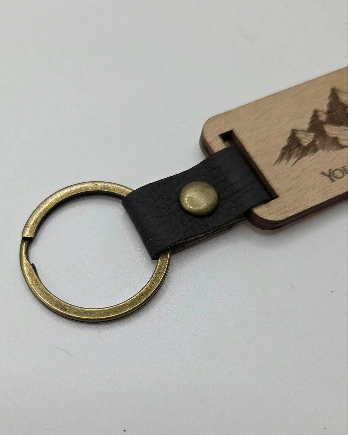 A close-up of a keychain with a split brass key ring, a black leather strap with a brass snap, and a light wood tag engraved with a mountain and pine tree design.