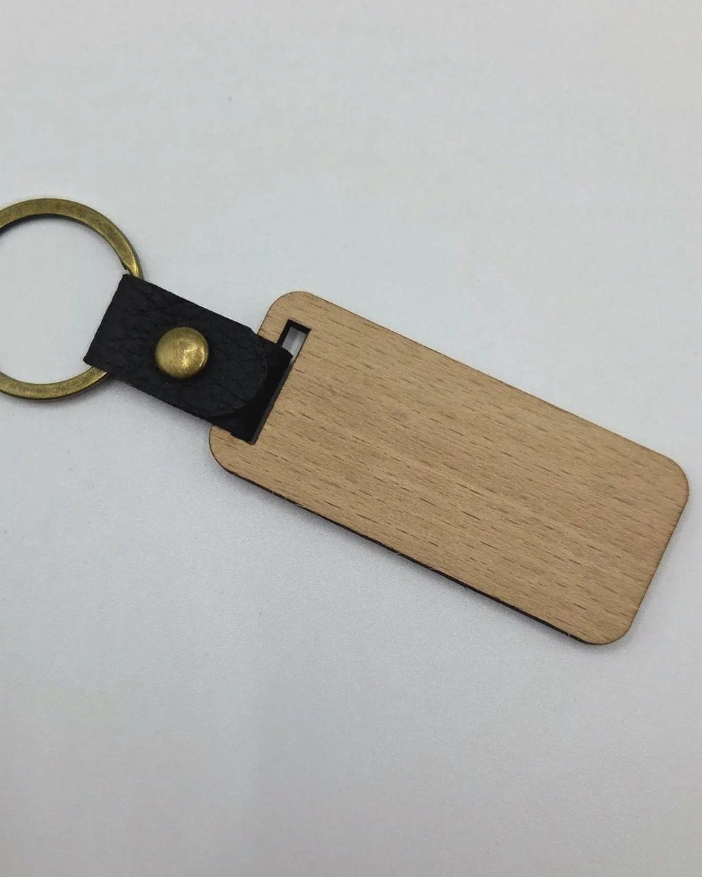 A blank wooden keychain tag with rounded corners, attached to a black leather loop with a brass rivet and a gold-tone split key ring, shown on a plain light background.
