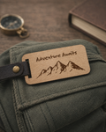 A rectangular wooden luggage tag engraved with 'Adventure Awaits' and a mountain illustration, attached to a black leather strap on an olive green canvas bag. A brass compass and a brown book are blurred in the background on a wooden surface.