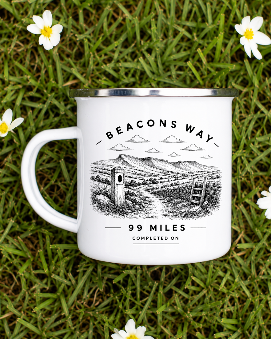 A white enamel camping mug with a silver rim, featuring a black and white illustration of a scenic road and the text 'BEACONS WAY 99 MILES COMPLETED ON', placed on green grass with small white daisies scattered around it.