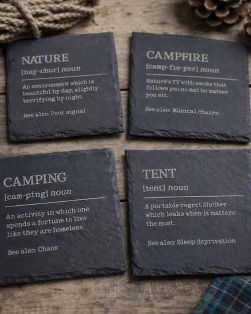 Four dark gray slate coasters with definitions of camping-related words on a wooden surface.
