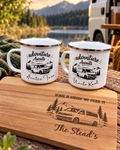 Two white enamel camp-style mugs with black camper illustrations and text reading 'Adventure Awaits' on a wooden engraved cutting board outdoors. The board reads 'Home is where we park it' and 'The Stead's'.