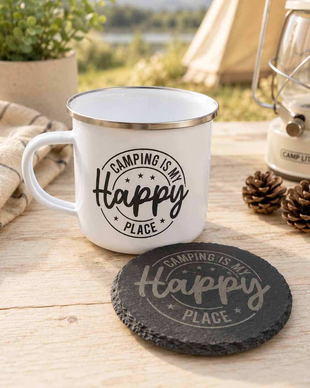 A white enamel camping mug with a silver rim sits on a wooden table outdoors, printed with the phrase 'CAMPING IS MY Happy PLACE.' A matching round dark slate coaster is in front.