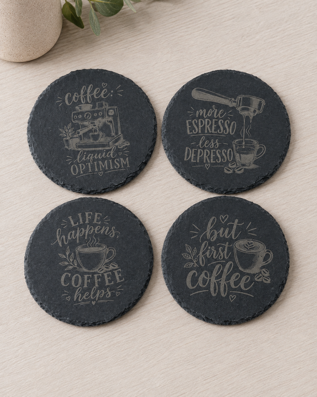 Four round dark slate coasters with coffee-themed quotes and illustrations, arranged on a light wood surface with a small vase in the corner.