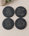 Four round dark slate coasters with coffee-themed quotes and illustrations, arranged on a light wood surface with a small vase in the corner.