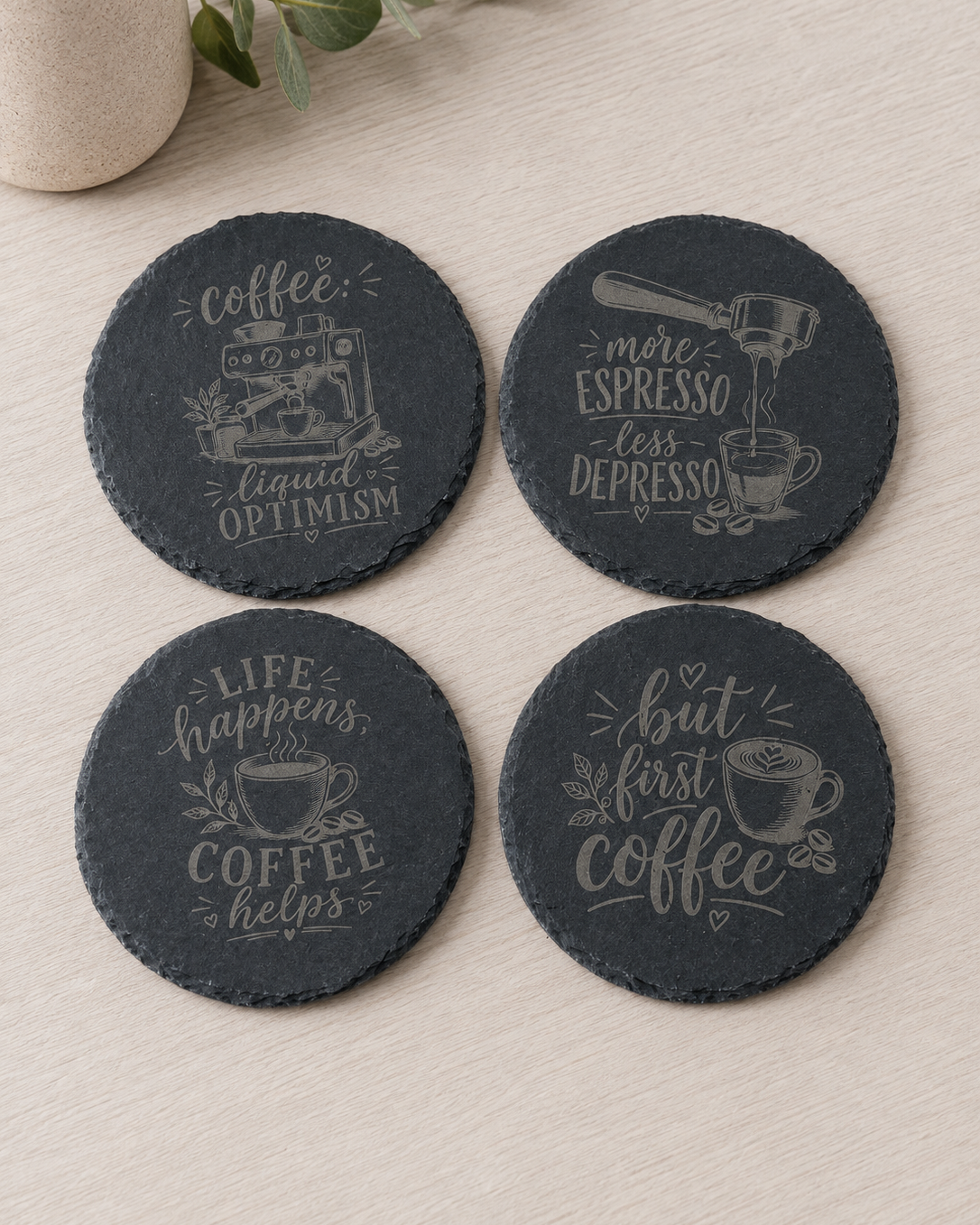 Four round dark slate coasters with coffee-themed quotes and illustrations, arranged on a light wood surface with a small vase in the corner.