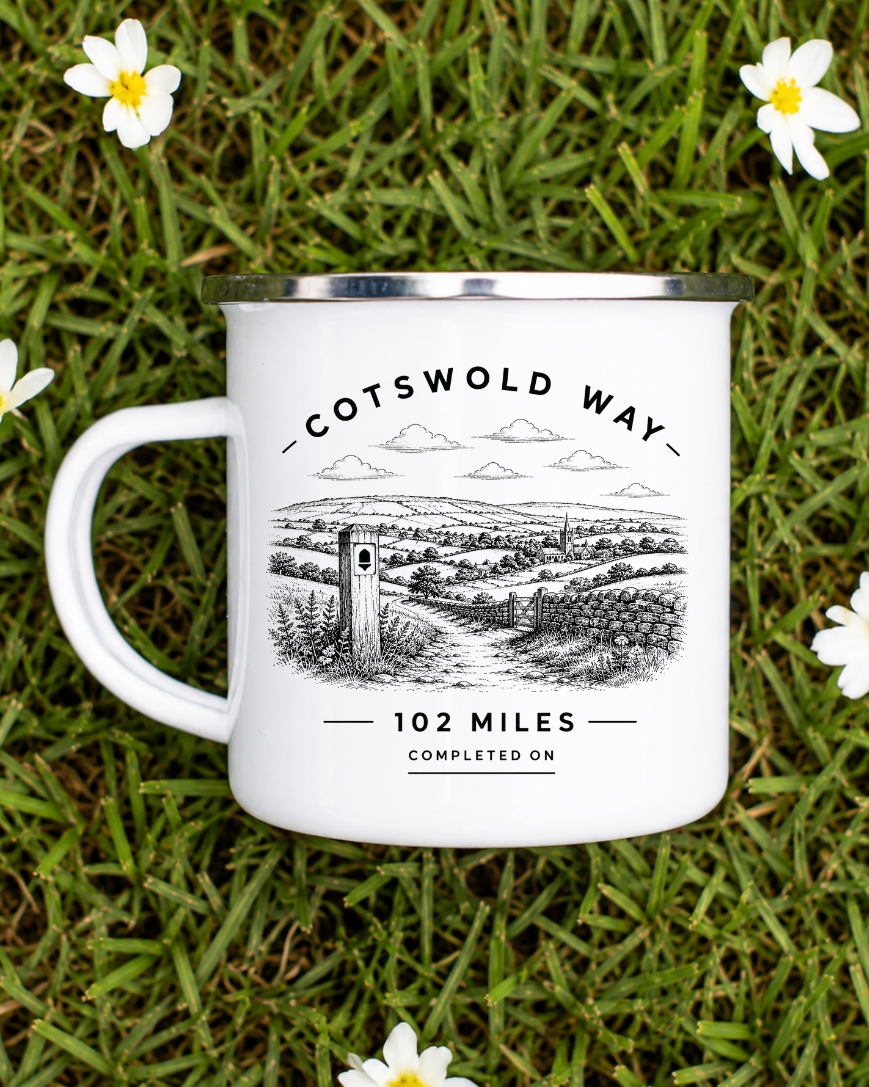 A white enamel-style camping mug with a silver rim, printed with the Cotswold Way trail illustration and text, placed on green grass with small white flowers around it.
