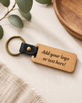 A personalized wooden keychain with a black leather loop and brass key ring, shown on a neutral textured surface with decorative eucalyptus leaves and a wooden plate.