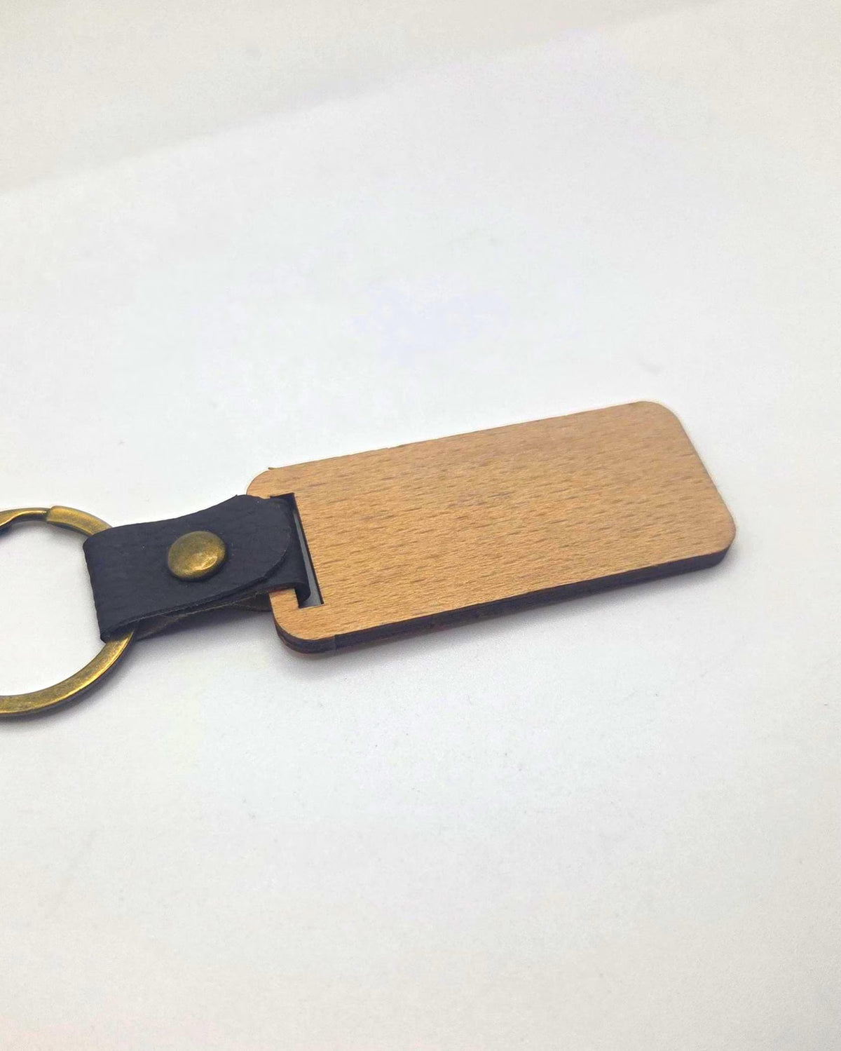 A blank wooden keychain tag with a light brown wood grain finish, attached to a black leather loop with a brass snap and a gold-tone split keyring, shown on a plain white background.