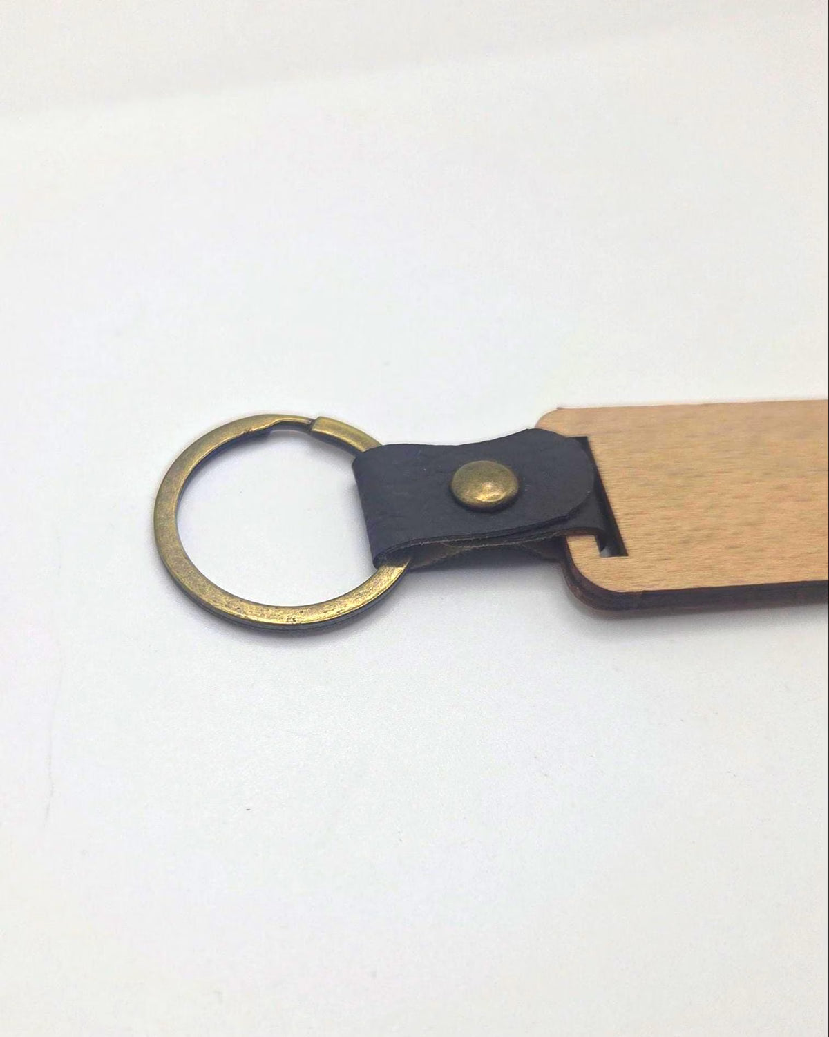 A close-up of a keychain or bag charm featuring a light wood tab, a black leather loop, and a brass split ring on a plain white background.