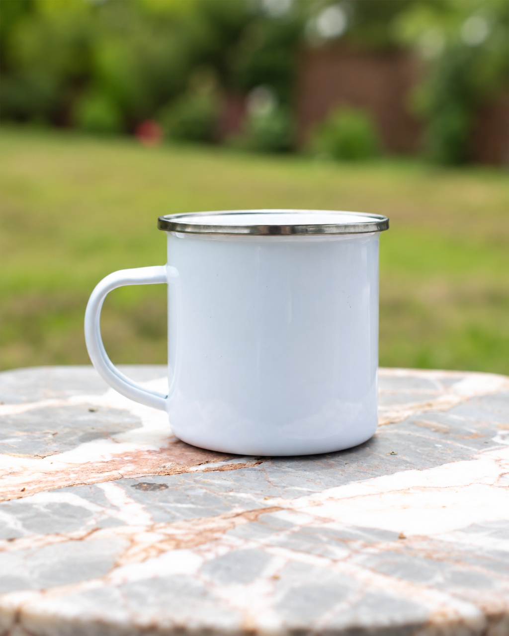 A white enamel camping mug with a silver metal rim and handle, sitting on a stone table with a blurred green garden background.