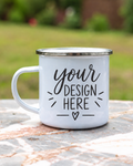 A white enamel camping mug with a silver metal rim and handle, featuring the customizable black text 'your DESIGN HERE' on the front, resting on a stone surface with a blurred green outdoor background.