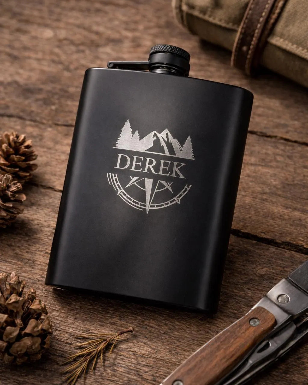 A matte black metal flask with a silver engraved design of mountains, trees, and the name 'DEREK' with flags, displayed on a rustic wooden surface with pinecones, a folding knife, and a canvas bag.
