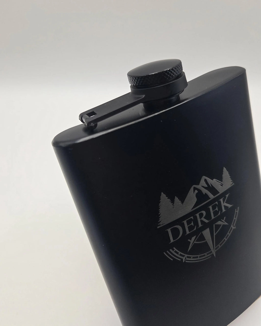 A matte black metal flask with a matching black screw cap and carrying clip, featuring a silver engraved logo of mountains, trees, and the name 'DEREK' on the front, set against a plain light gray background.