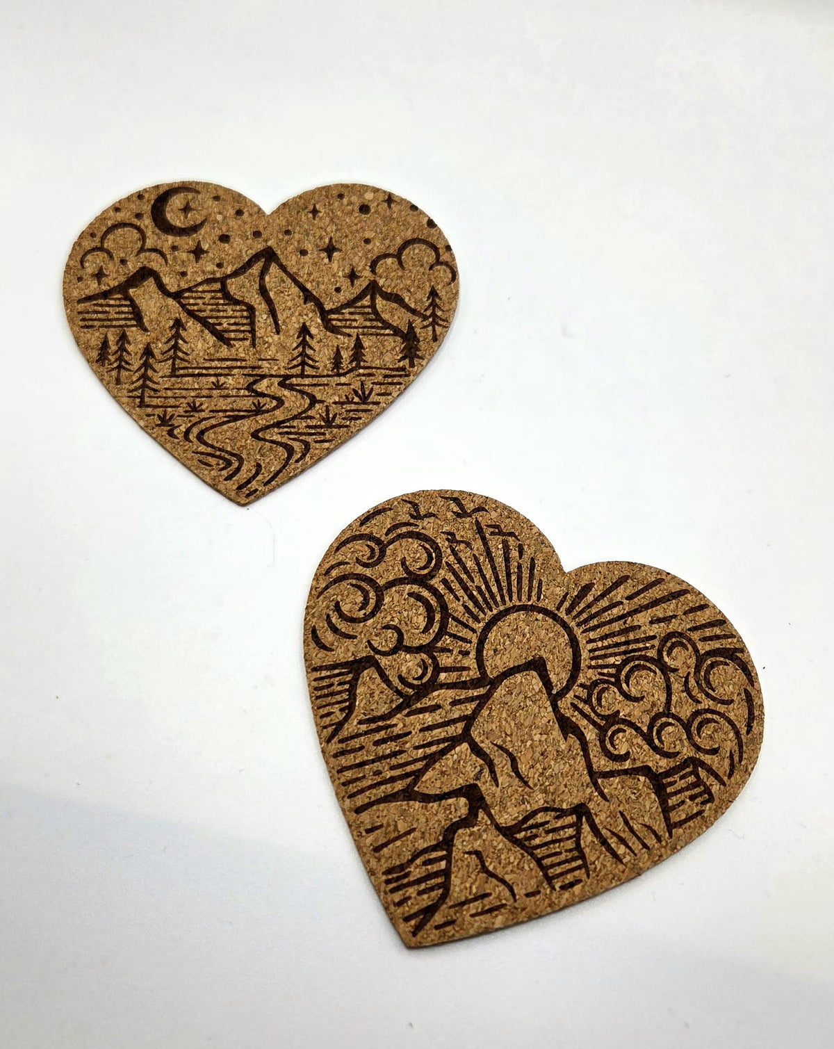 Two heart-shaped cork coasters with engraved nature designs, one showing a mountain landscape under a crescent moon and stars, and the other showing a mountain scene with a sun and clouds, on a plain white background.