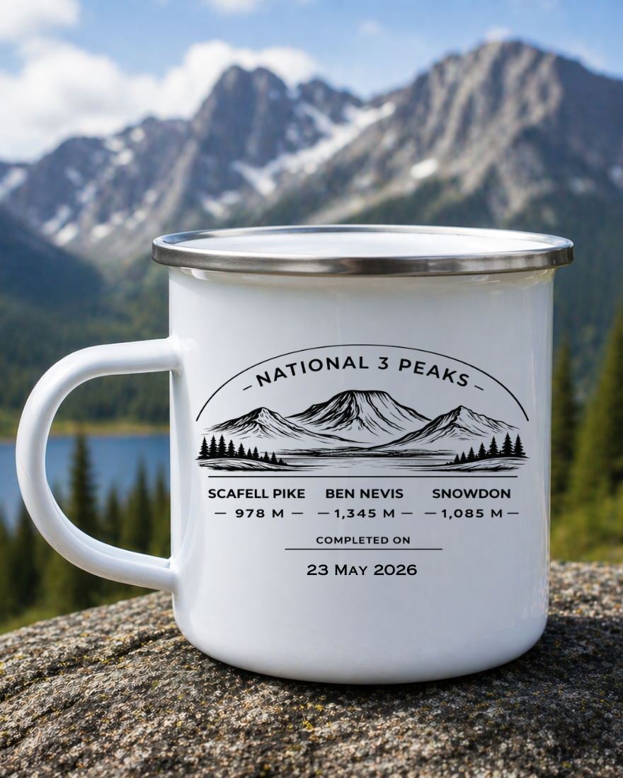 A white enamel camping mug with a silver rim sits on a rock in front of a mountain landscape. The mug is printed with a black mountain graphic and the text 'National 3 Peaks' along with elevation names and dates for Scafell Pike, Ben Nevis, and Snowdon.