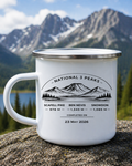 A white enamel camping mug with a silver rim sits on a rock in front of a mountain landscape. The mug is printed with a black mountain graphic and the text 'National 3 Peaks' along with elevation names and dates for Scafell Pike, Ben Nevis, and Snowdon.