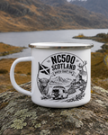 A white enamel camping mug with a silver rim and black graphic print reading 'NC500 SCOTLAND NORTH COAST' with a van, mountains, and deer, placed on a rock with a scenic mountain lake background.