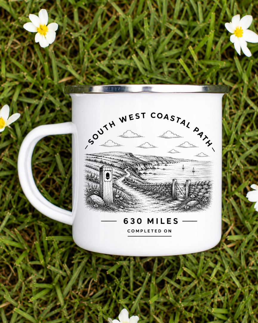 A white enamel-style mug with a silver rim and handle, printed with a black-and-white coastal path illustration and the text 'SOUTH WEST COASTAL PATH', '630 MILES', and 'COMPLETED ON'. The mug is placed on green grass surrounded by small white flowers.