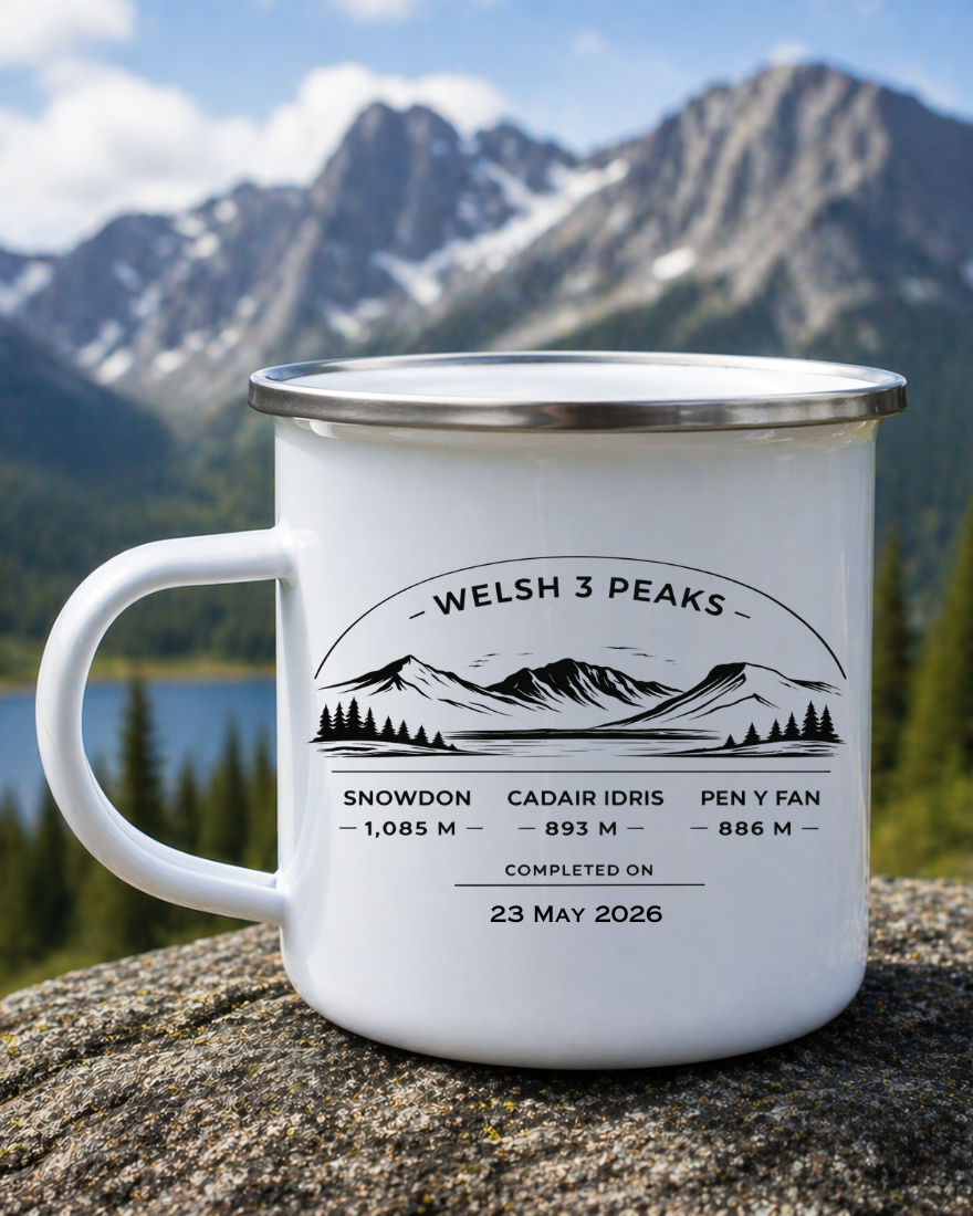 A white enamel-style mug with a silver rim and handle, printed with a black mountain graphic and the text 'WELSH 3 PEAKS' and summit elevations, photographed outdoors on a rock with mountains and trees in the background.