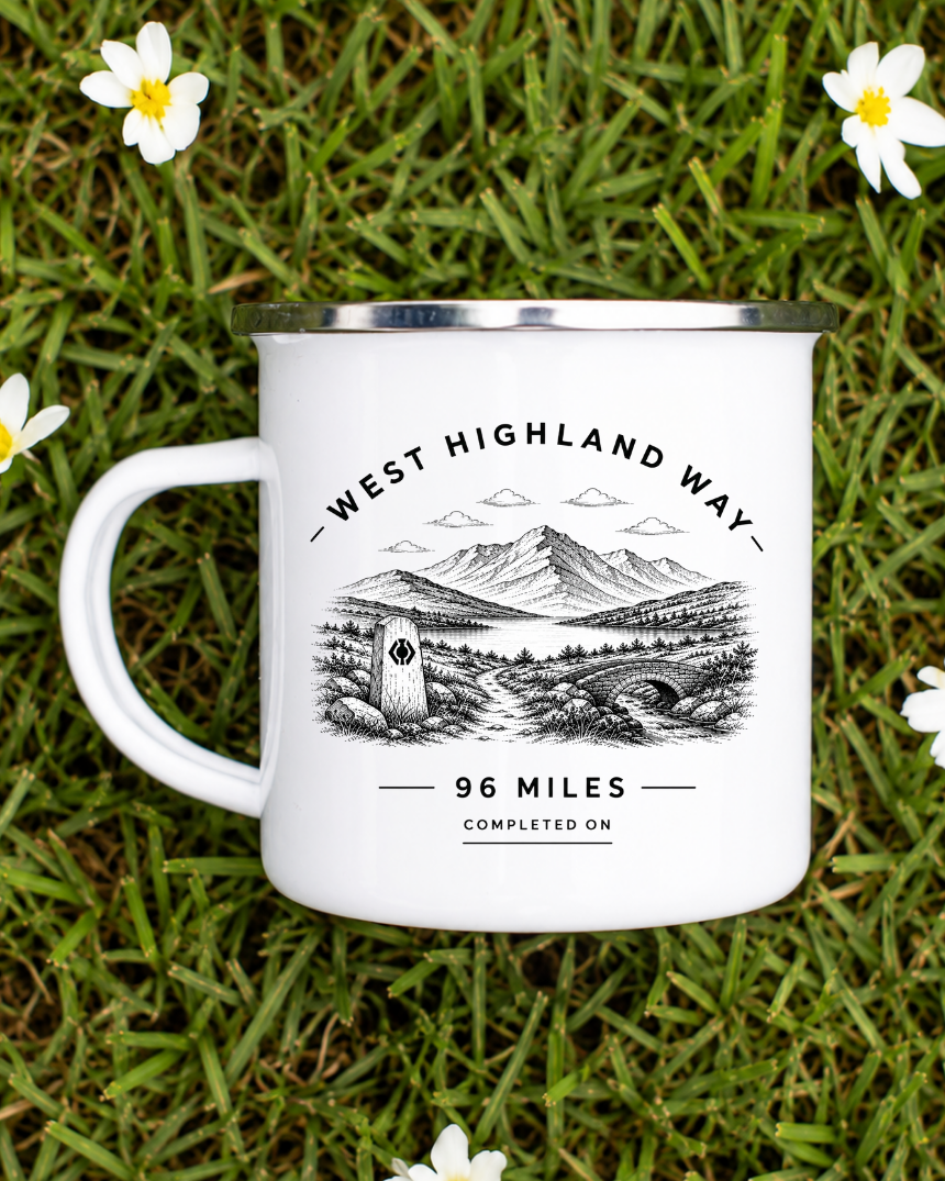 A white enamel-style camping mug with a silver rim sits on green grass surrounded by small white daisies. The mug has a black illustrated landscape print and text reading “WEST HIGHLAND WAY” and “96 MILES COMPLETED ON.