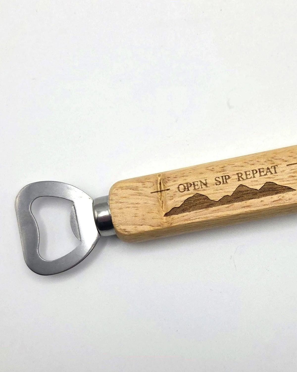 A bottle opener with a natural wood handle engraved with 'OPEN SIP REPEAT' and a mountain design, paired with a silver metal blade, shown on a plain white background.