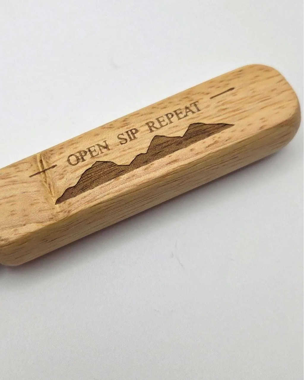 A close-up of a bottle opener with a light natural wood handle engraved with the words 'OPEN SIP REPEAT' and a mountain silhouette, with a silver metal blade partially visible on the left, set against a plain light gray background.