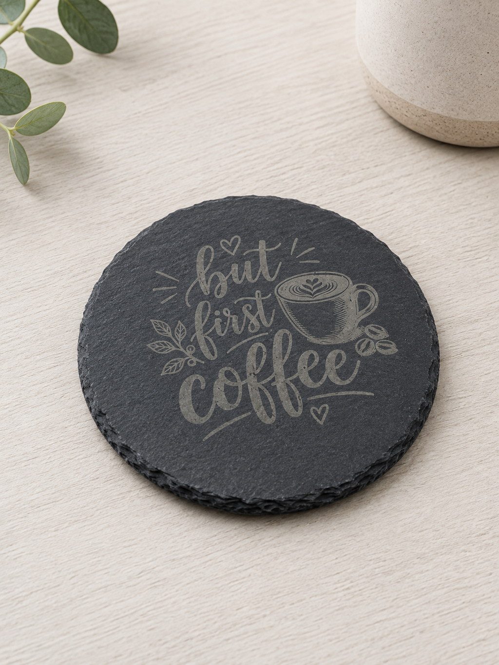 A round black slate coaster with a light gray engraved design that reads 'but first coffee' with a coffee cup illustration, placed on a light wood surface.