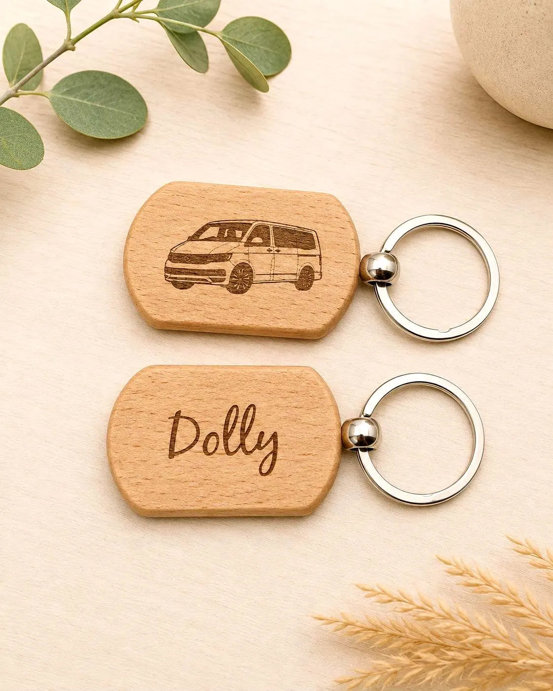 Two personalized wooden keychains with silver key rings, displayed on a light beige background with decorative green leaves and wheat. One keychain features an engraved van, and the other is personalized with the name 'Dolly'.