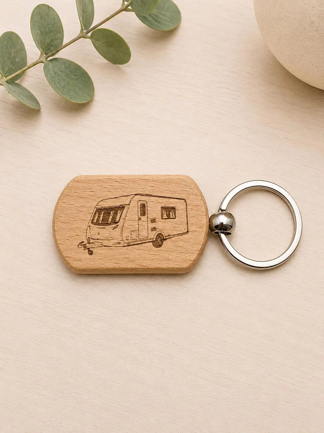 A wooden keychain with an engraved camper trailer design, attached to a silver metal key ring, displayed on a light beige wooden surface with decorative green leaves and a stone.