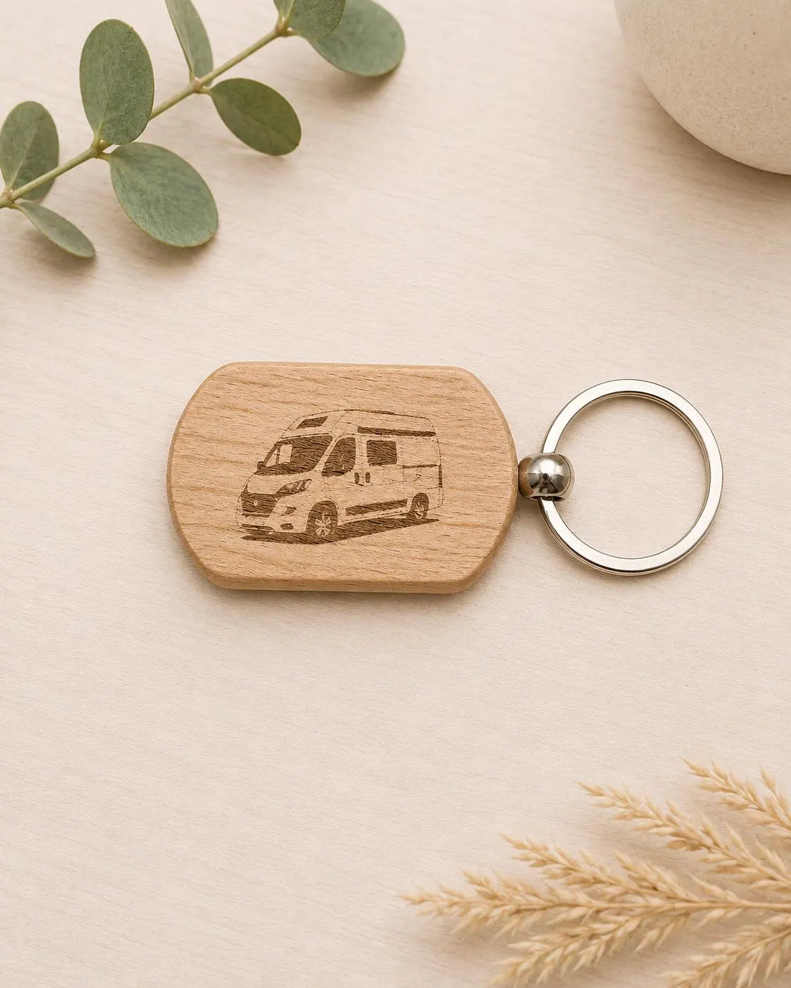 A wooden keychain with an engraved camper van design, attached to a silver metal key ring, displayed on a light beige wooden surface with decorative eucalyptus leaves and dried wheat.