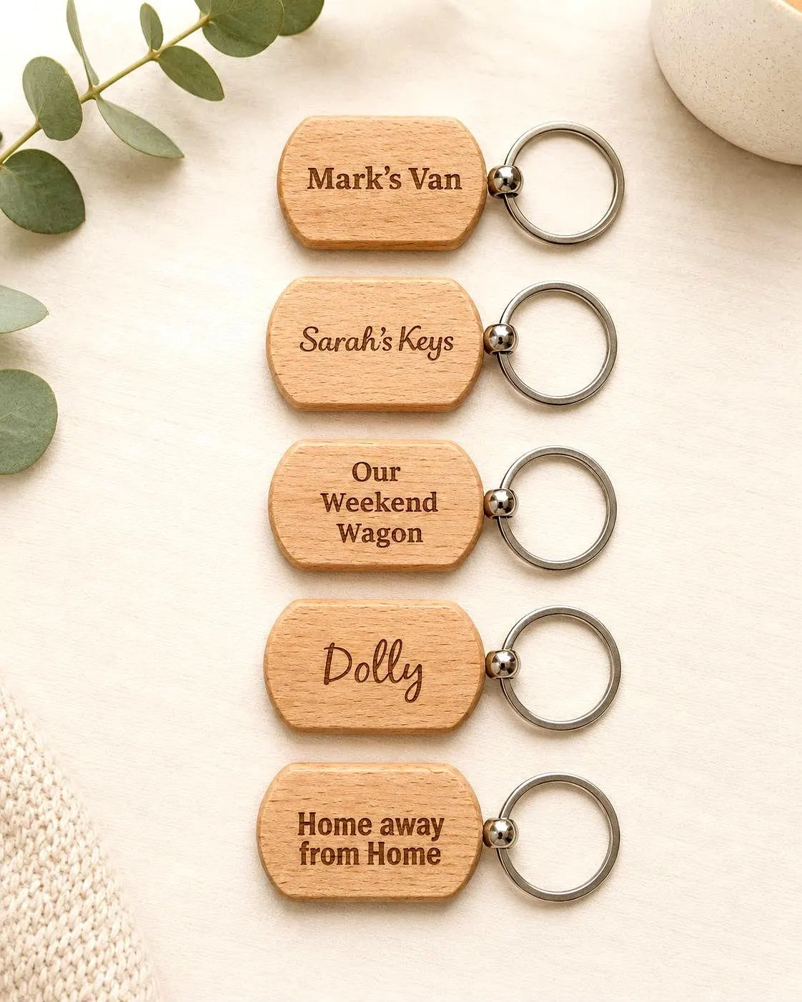 Five personalized wooden keychains with silver metal key rings, arranged vertically on a light beige textured background with decorative eucalyptus leaves and a stone.