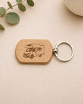 A wooden dog-tag style keychain with an engraved camper van design, attached to a silver metal key ring, photographed on a light wood surface with a eucalyptus sprig and part of a ceramic object in the background.