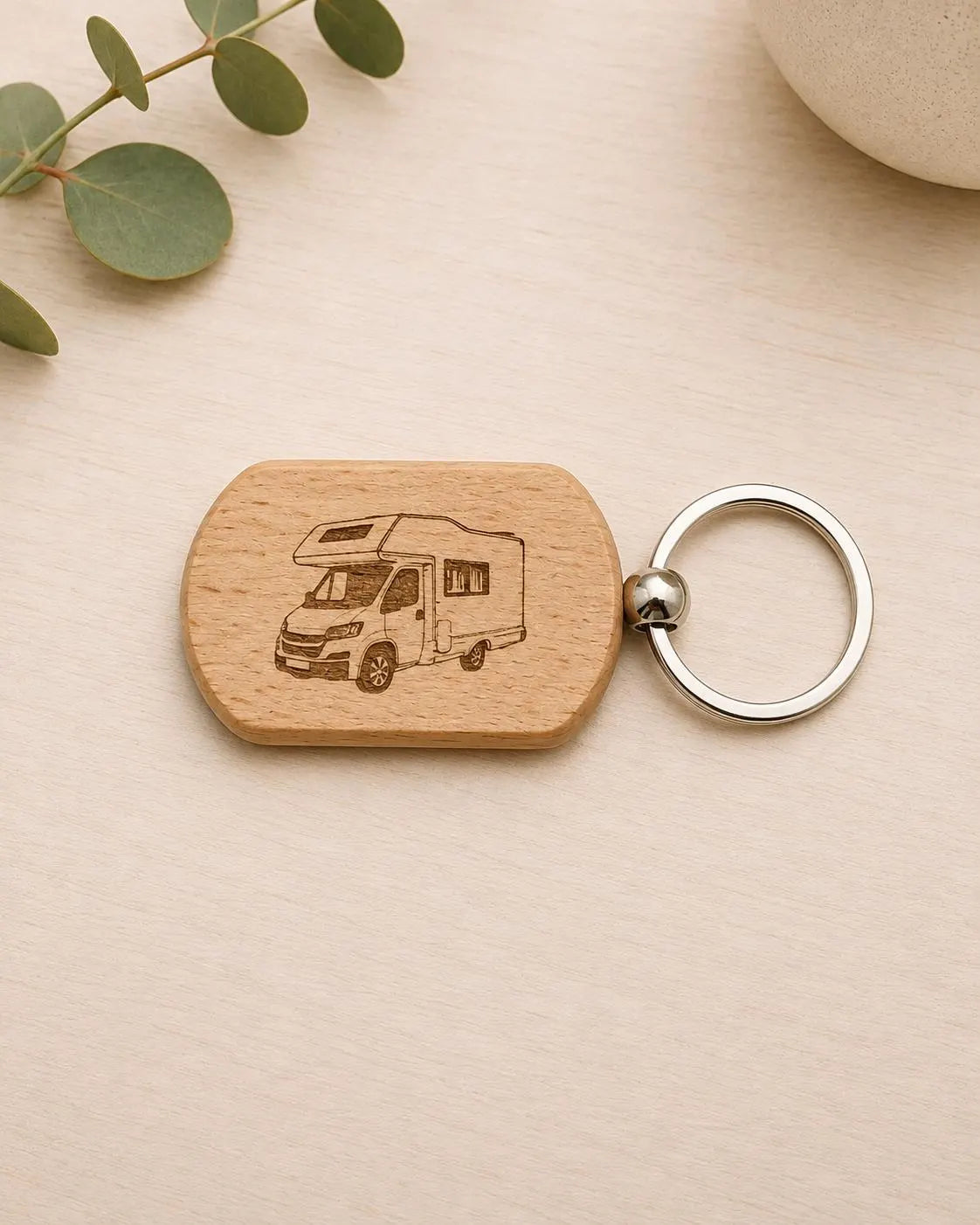 A wooden dog-tag style keychain with an engraved camper van design, attached to a silver metal key ring, photographed on a light wood surface with a eucalyptus sprig and part of a ceramic object in the background.
