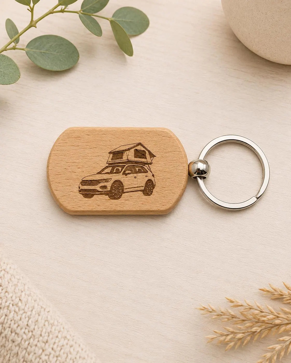 A wooden keychain with a rounded rectangular tag engraved with a car with a rooftop tent, attached to a silver metal key ring, displayed on a light wood surface with decorative eucalyptus leaves and a knit fabric.