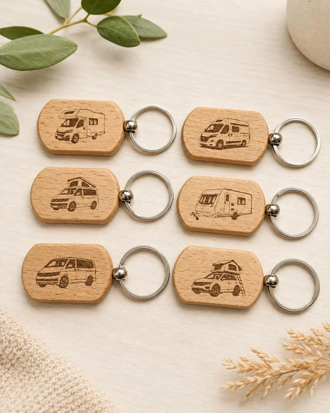 Six personalized wooden keychains with engraved camper van illustrations and silver metal key rings, arranged on a light beige textured surface with decorative leaves and dried grass.