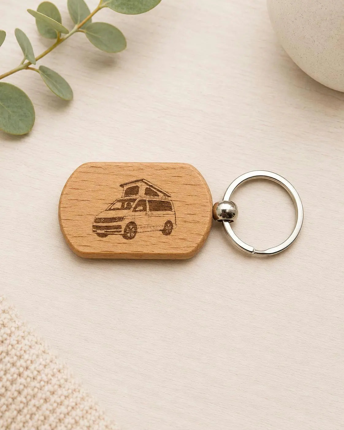 A wooden keychain with a rounded rectangular block engraved with a vintage camper van, attached to a silver split ring, displayed on a light beige textured surface with eucalyptus leaves.