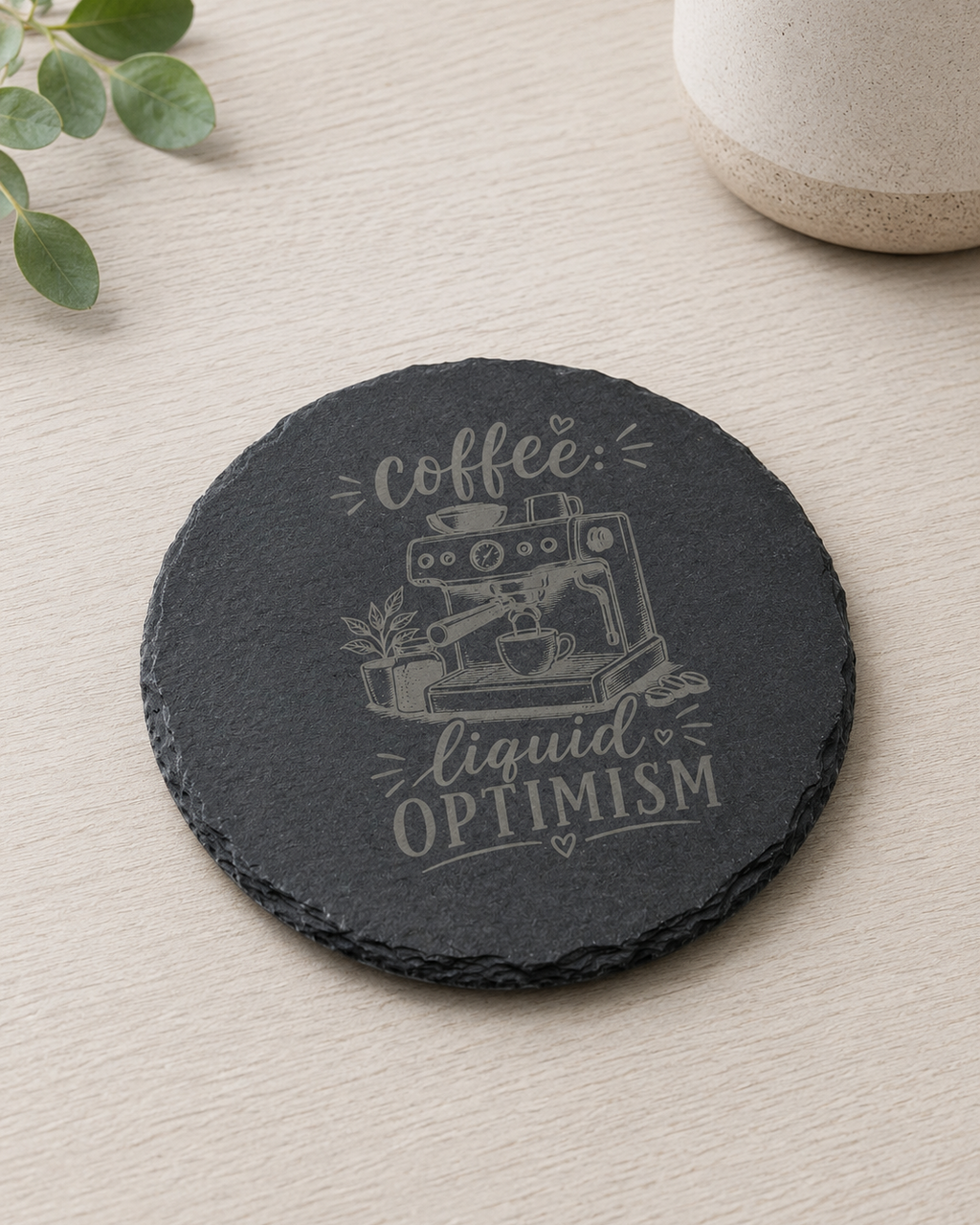 A round black slate coaster with a light gray engraved design featuring an espresso machine and the words 'coffee liquid OPTIMISM', shown on a light wood tabletop with a ceramic cup and green leaves as props.