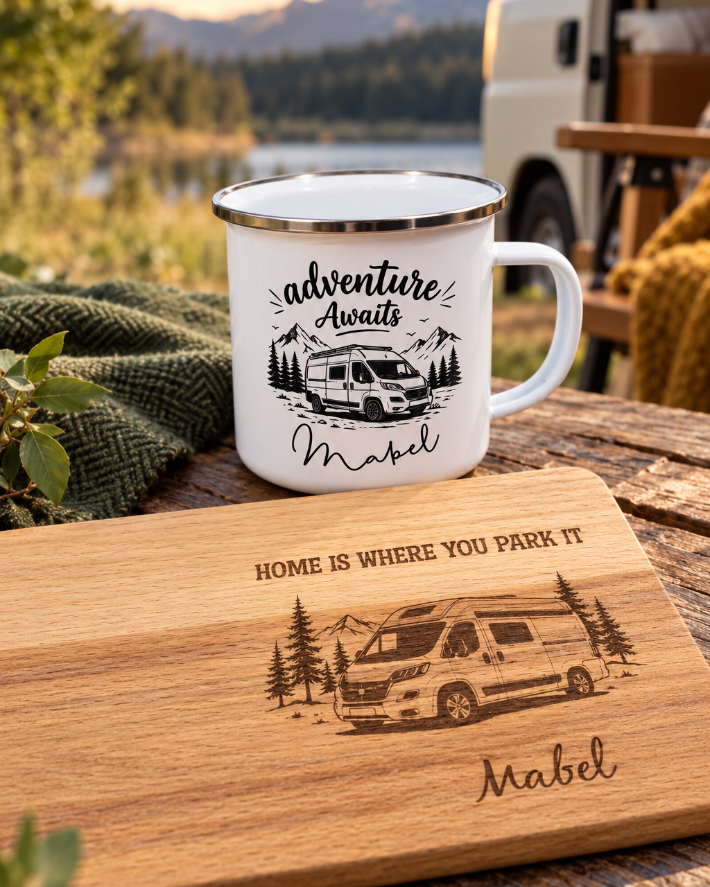 A white enamel camping mug with a black graphic of a camper van and the text 'Adventure Awaits Mabel' sits on a wooden table outdoors. In front of it is a wooden cutting board engraved with the phrase 'Home is where you park it' and a matching camper van illustration with the name 'Mabel'.