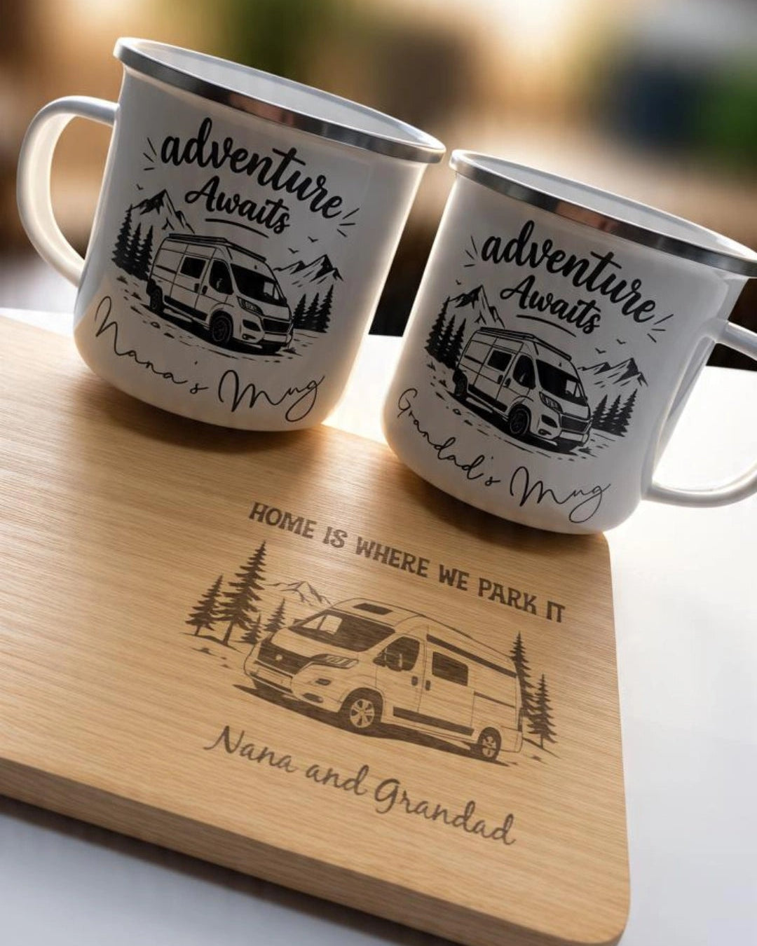 Two white enamel campfire mugs with black engraved camper illustrations and the words 'Adventure Awaits' and 'Nana & Grandad' sit on a wooden engraved tray. The tray also features a campervan illustration and the phrase 'Home is where we park it.