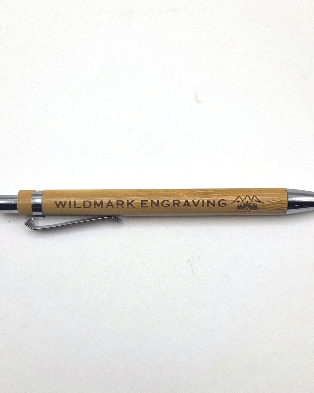 A wooden pen with silver metal accents, engraved with 'WILDMARK ENGRAVING' and a mountain logo, resting on a plain white surface.
