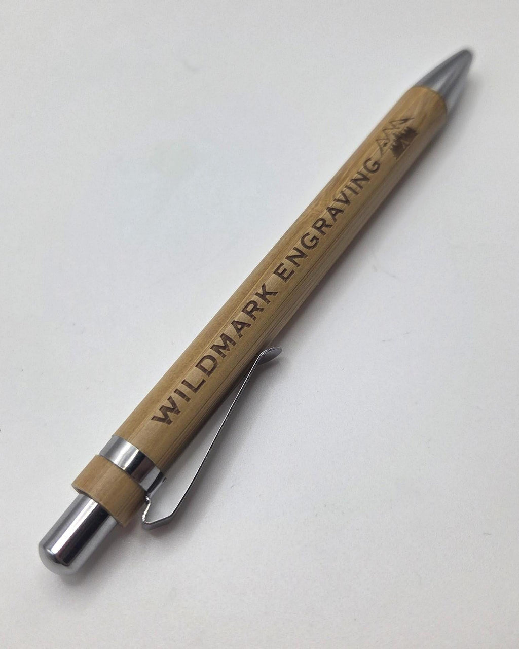 A wooden pen engraved with 'WILDMARK ENGRAVING' and a deer logo, featuring a silver metal clip and tip on a light gray background.
