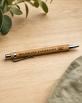 A wooden pen with silver metal accents engraved with 'WILDMARK ENGRAVING' and a mountain logo, resting on a light wood tabletop with green leaves and a textured fabric in the background.