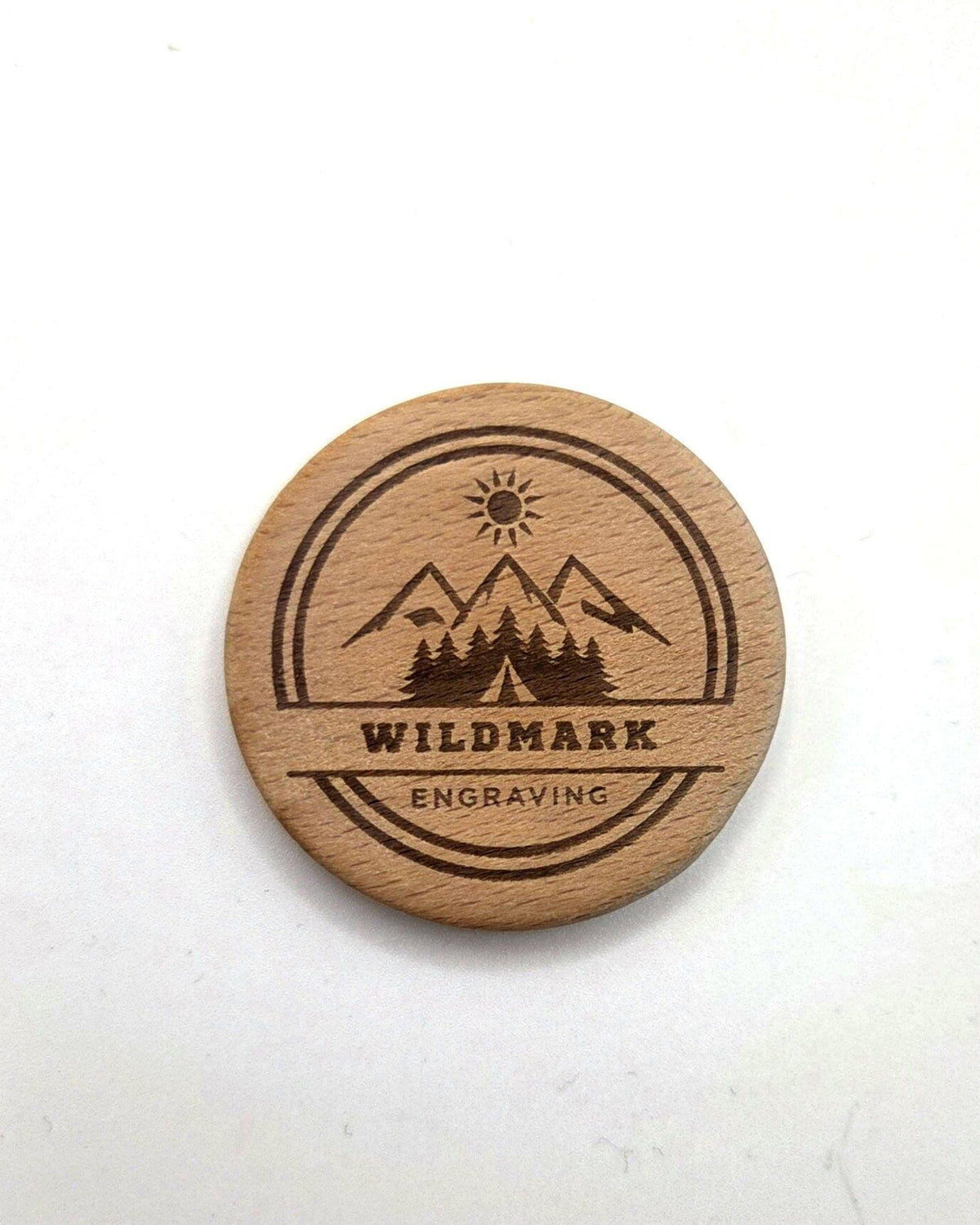 A round natural wood coaster engraved with a mountain, sun, and trees design, featuring the text 'WILDMARK ENGRAVING' in the center, set against a plain off-white background.
