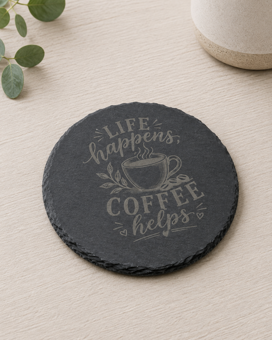 A round black slate coaster with a light gray engraved design featuring a coffee cup and the phrase 'Life happens, coffee helps', displayed on a light wood tabletop with a ceramic mug and green leaves in the background.